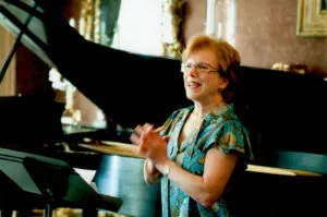 Mary Falvey speaking to an audience beside a grand piano in an ornate room